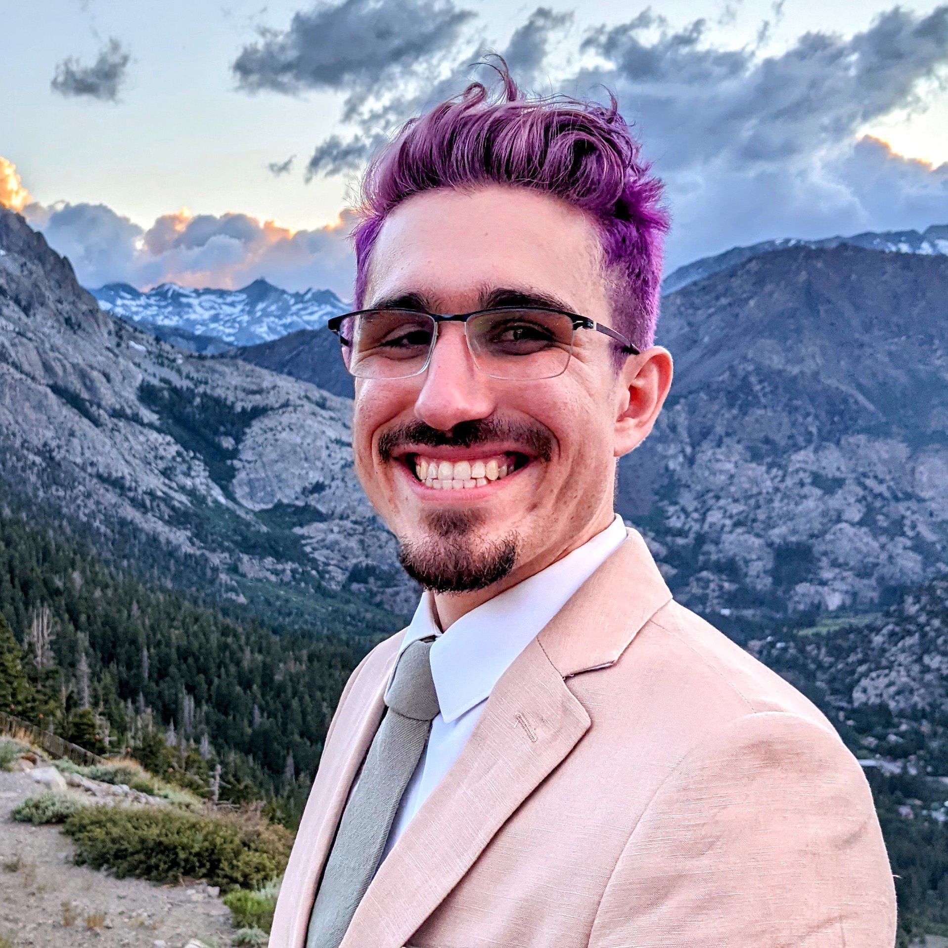 Dani in front of a mountain range in a suit and tie. They have purple hair and slightly tinted glasses.